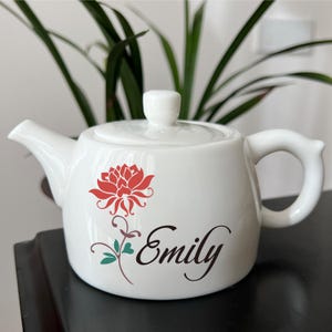 May include: White ceramic teapot with a red floral design and the name "Emily" in script. The teapot has a lid, spout, and handle. A green plant is in the background.