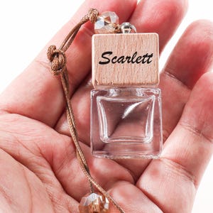 Pode incluir: Uma pequena garrafa de vidro transparente com uma tampa de madeira, com o nome "Scarlett" em escrita preta. A garrafa é suspensa por um cordão castanho com contas de cristal. A garrafa é segurada em uma mão.