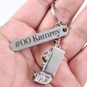 Puede incluir: Un llavero plateado con un diseño de camión en miniatura. El llavero incluye una etiqueta rectangular grabada con el texto "#00 Kamrey". El camión tiene detalles, incluyendo ruedas y un remolque.