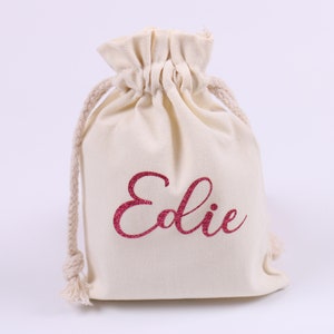 May include: A white cotton drawstring bag with the name "Edie" printed in pink glitter.