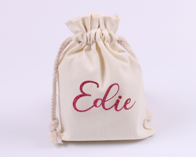 May include: A white cotton drawstring bag with the name "Edie" printed in pink glitter.
