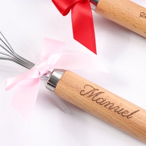 May include: A wooden whisk with a pink ribbon tied around the handle. The handle is engraved with the name "Manuel".