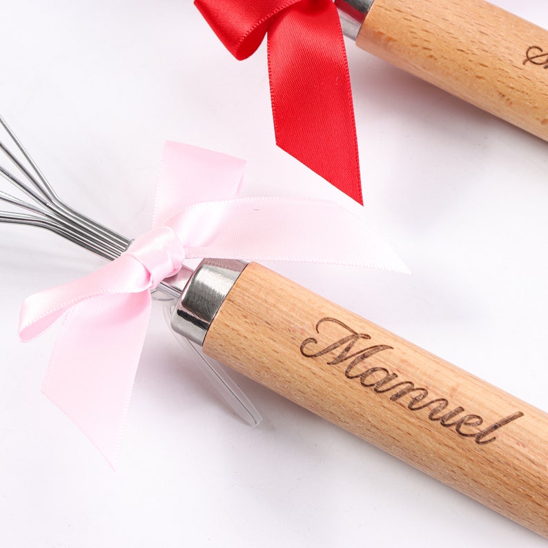 May include: A wooden whisk with a pink ribbon tied around the handle. The handle is engraved with the name "Manuel".