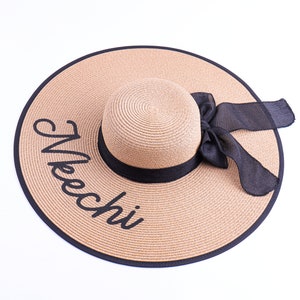 May include: A wide-brimmed straw sun hat with a black ribbon and bow. The hat has the word "Beach" embroidered on the brim in black.