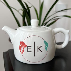 Op de afbeelding: Witte keramische theepot met deksel en handvat. De theepot heeft een bloemenontwerp met de letters "EK" in een cirkelvormig frame. Een rode bloem en een turquoise blad accentueren het ontwerp. De theepot staat op een zwart oppervlak.