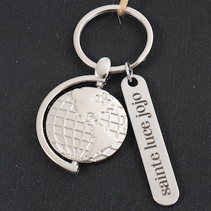 May include: A silver-coloured keyring with a globe design and a rectangular tag. The globe shows continents and grid lines. The tag reads "sainte luce jojo" vertically. A silver ring and a brown string are also visible.