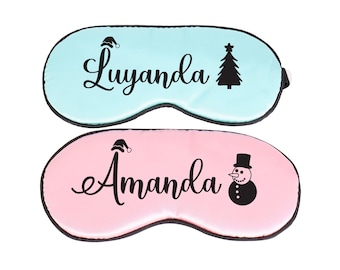 Op de afbeelding: Twee slaapmaskers, één lichtblauw en één roze, met zwarte rand. Het lichtblauwe masker heeft de naam "Luyanda" en een kerstboomafbeelding. Het roze masker heeft de naam "Amanda" en een sneeuwpopafbeelding.