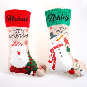 Può includere: Due calze di Natale con polsini rossi e verdi. Una calza presenta un disegno di Babbo Natale, l'altra un pupazzo di neve. Entrambe le calze hanno le parole "Buon Natale" e un nome ricamato.