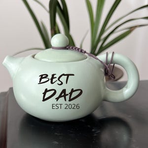 May include: A light green ceramic teapot with a lid and handle. The teapot has the words "BEST DAD EST 2026" printed on the front in black. A small, round knob sits atop the lid, and a decorative cord is attached.