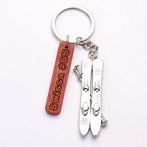 Op de afbeelding: Een houten sleutelhanger met de tekst "Swissa #55" en een zilveren metalen hanger met een paar ski's met stokken.