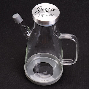 May include: Clear glass oil dispenser with a gray base and handle. The lid is silver with the name "Jessie" and the date "July 12, 2025" engraved on it. The dispenser has measurement markings from 100ml to 500ml.
