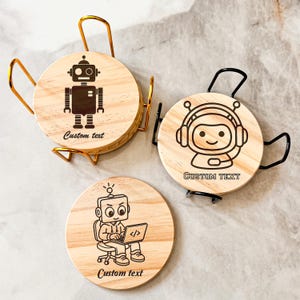 May include: Three round wooden coasters featuring black laser-engraved robot illustrations and the text Custom text. Two coasters are held in metal wire holders, one gold and one black, while the third coaster sits flat with a coder robot design.
