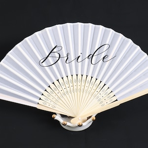 May include: A white and light gray folding fan with the word "Bride" in elegant script. The fan has a cream-colored wooden handle and is displayed on a white decorative stand. The fan is open, showcasing its pleated design.