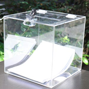 May include: A clear acrylic ballot box with a slot on top and a hinged, lockable door. The box contains a stack of white papers. A set of keys is in the lock. The box is on a dark surface with a blurred green background.