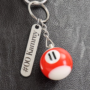 May include: A silver keychain with a rectangular tag engraved with "#00 Kamrey" and a red and white billiard ball charm. The billiard ball is number 11.