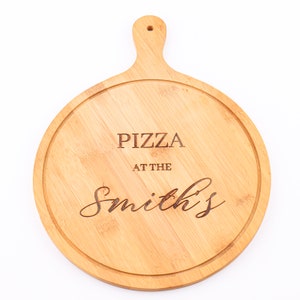 May include: Round wooden pizza serving board with a handle. The board has a light brown color and features the engraved text "PIZZA AT THE Smith's". The board has a groove around the edge to prevent spills.