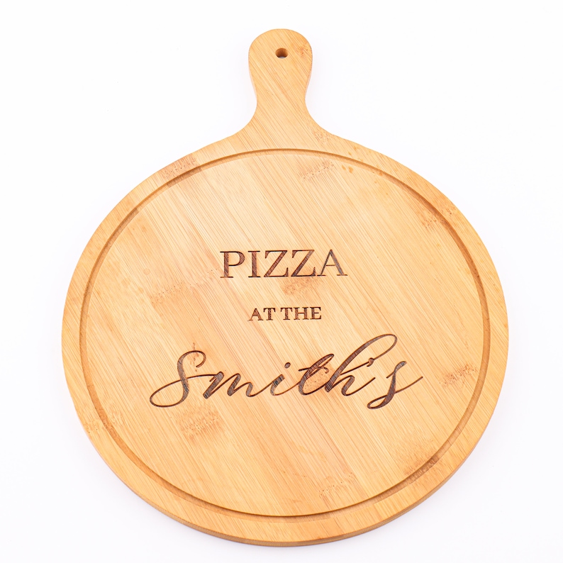 May include: Round wooden pizza serving board with a handle. The board has a light brown color and features the engraved text "PIZZA AT THE Smith's". The board has a groove around the edge to prevent spills.