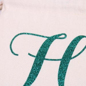 May include: A white canvas bag with a teal glitter letter "H" in a cursive font.