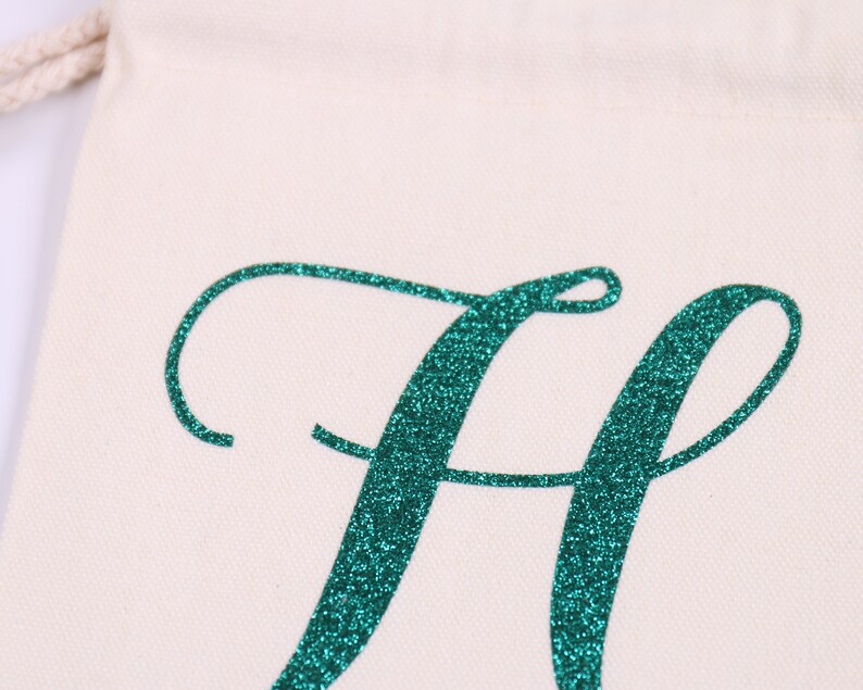 May include: A white canvas bag with a teal glitter letter "H" in a cursive font.