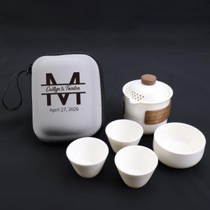 Puede incluir: Un juego de té blanco con un estuche. El juego incluye una tetera con asa de madera, cuatro tazas de té pequeñas y un estuche blanco con el texto "Caitlyn & Trenton" y la fecha "27 de abril de 2026".