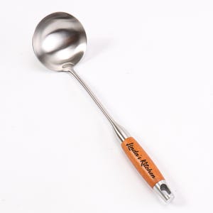 Op de afbeelding: Een zilveren soeplepel met een houten handvat. Het handvat heeft de tekst "Linda's Kitchen" gegraveerd.
