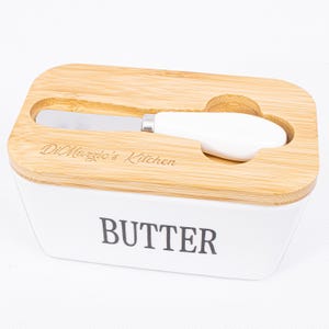 Könnte beinhalten: Eine weiße Keramik-Butterdose mit einem Holzdeckel. Der Deckel hat einen Schlitz für ein Buttermesser und ist mit dem Text "DiMeglio's Kitchen" graviert. Die Dose hat das Wort "BUTTER" auf der Vorderseite.