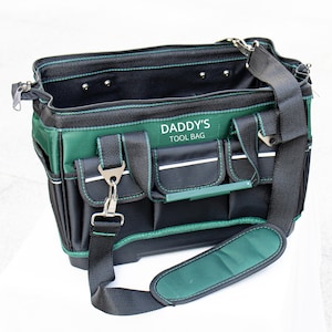 May include: A black and dark green tool bag featuring multiple exterior pockets, a wide open top, and a detachable shoulder strap. The front of the bag is embroidered with the white text DADDY'S TOOL BAG. The bag includes metal hardware.