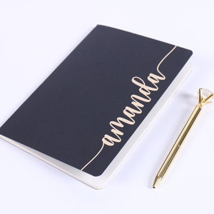 May include: A dark blue notebook with the name "Amanda" written in gold script on the cover. A gold pen with a clear diamond-shaped top is next to the notebook.