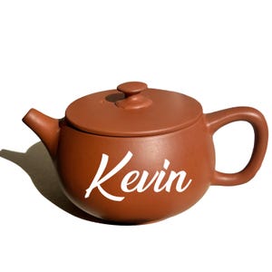 May include: A brown ceramic teapot with a lid and handle. The teapot has the name "Kevin" in white script on the side. The teapot is designed for brewing tea.