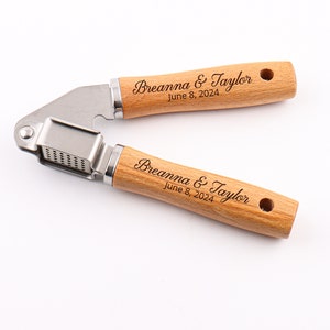 May include: A wooden garlic press with a stainless steel crusher. The handle is engraved with the names "Breanna & Taylor" and the date "June 8, 2024".