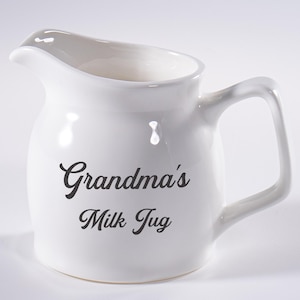 Puede incluir: Jarra de leche de cerámica blanca con un pico curvo y un asa. Las palabras "Grandma's Milk Jug" están impresas en negro en la parte delantera. La jarra tiene un estilo clásico y rústico.
