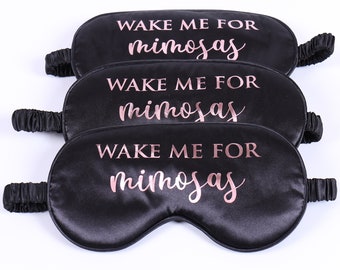 Op de afbeelding: Drie zwarte satijnen slaapmaskers met de tekst "Wake Me For Mimosas" gedrukt in roségouden letters.