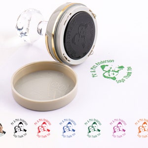 Op de afbeelding: Een zelf-inktende stempel met een transparante acrylhandgreep en een rond inktkussen. Het inktkussen is zwart en heeft een ontwerp van een stel dat hand in hand staat. De stempel wordt gebruikt om een ​​groene afdruk te maken die "Mr & Mrs Anderson Says Thank You" zegt.