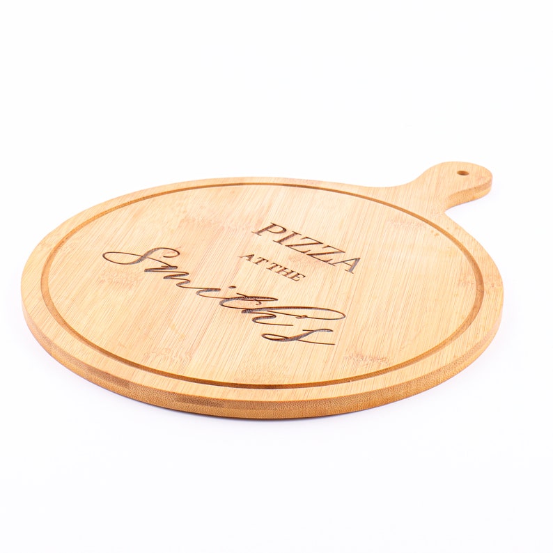 May include: A round, light brown bamboo pizza serving board with a handle. The board has a carved groove around the edge and the words "PIZZA AT THE Smith's" are etched into the wood.