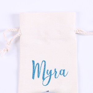 May include: A white cotton drawstring bag with a blue glitter name "Myra" printed on the front.