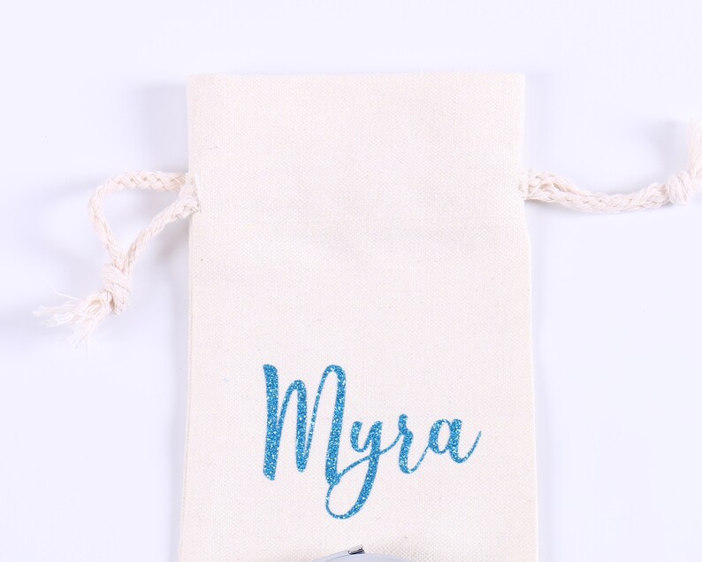 May include: A white cotton drawstring bag with a blue glitter name "Myra" printed on the front.