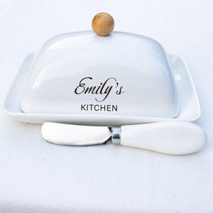 Puede incluir: Una mantequillera de cerámica blanca con base rectangular y tapa abovedada que incluye un pomo de madera redondo. La tapa tiene impreso el texto negro Emily's KITCHEN. Se incluye un cuchillo para mantequilla a juego con mango de cerámica.