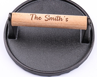 May include: A black cast iron burger press with a wooden handle. The handle is engraved with the text "The Smith's".