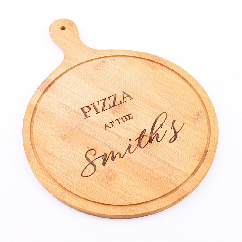 May include: A round, light brown bamboo pizza serving board with a handle. The board features a carved groove around the edge and the words "PIZZA at the Smith's" engraved in the center. The handle has a hole for hanging.
