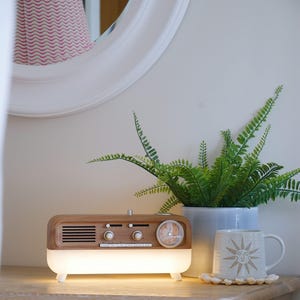 May include: A rectangular wooden radio with a built-in light base, featuring a speaker grille, two control knobs, several buttons, and an analog clock face on the right side. The radio emits a warm glow from its frosted white lower section.