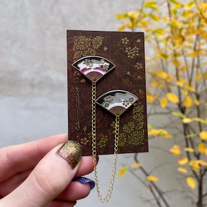 May include: Two fan-shaped enamel pins connected by a gold chain, displayed on a brown card. The pins have black, white, grey, and magenta designs. The card features a floral pattern and the word "KABUKI" printed vertically.