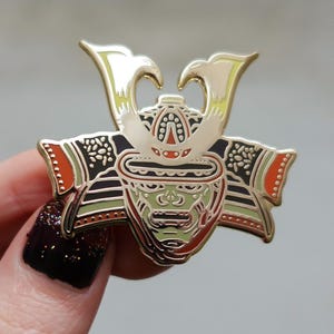 Puede incluir: Un pin de esmalte dorado con forma de casco de samurái. El pin presenta detalles intrincados con acentos rojos, verdes, negros y blancos. El casco tiene una cresta distintiva y elementos decorativos.