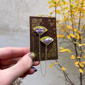 Può includere: Due spille a forma di ventaglio in smalto con catene dorate, attaccate a una carta marrone con motivi floreali. Le spille presentano una combinazione di colori giallo e viola con dettagli floreali. La carta ha la parola "KABUIRO" stampata verticalmente.