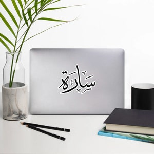 Könnte beinhalten: Ein schwarz-weißer Aufkleber mit dem arabischen Wort "Salat" in Kalligraphie. Der Aufkleber befindet sich auf einem silbernen Laptop.