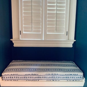 May include: A white window with white wooden shutters is above a white window seat with a blue and white patterned cushion.