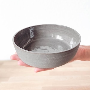 Könnte beinhalten: Eine handgefertigte, graue Keramikschale mit strukturierter Oberfläche. Die Schale ist rund mit einem leicht unregelmäßigen Rand und wird in einer Hand gehalten. Geeignet zum Servieren von Speisen.