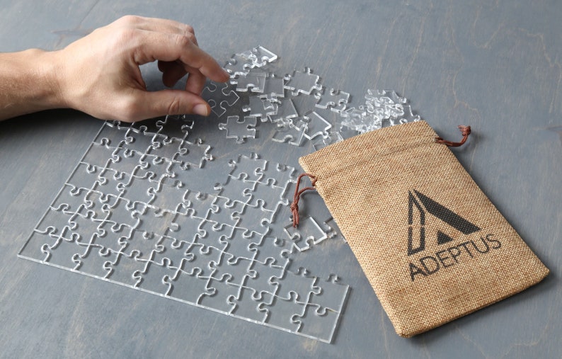 May include: A clear acrylic jigsaw puzzle with some pieces missing, being assembled on a gray surface. A burlap bag with a brown drawstring and the logo "ADEPTOS" printed on it is to the right of the puzzle.