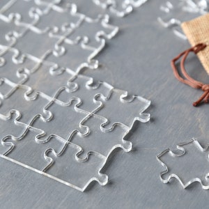 May include: A clear acrylic jigsaw puzzle with some pieces missing. The puzzle is on a gray surface and there is a brown burlap bag in the background.
