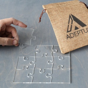 May include: A clear acrylic puzzle with six pieces, one piece is being held by a hand. The puzzle is on a gray surface. A burlap bag with the text "ADEPTUS" is in the background.
