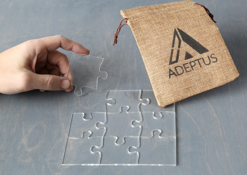 May include: A clear acrylic puzzle with six pieces, one piece is being held by a hand. The puzzle is on a gray surface. A burlap bag with the text "ADEPTUS" is in the background.